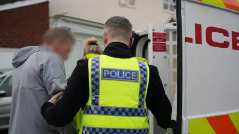 Two police officers are arresting someone (whose face is blurred) and loading them into a police van.