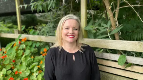 Leigh Yates looking at the camera and smiling. She is wearing a black top. Behind her are bushes with orange flowers and a wood fence.