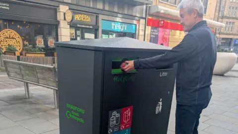 Man putting a red Coca-Cola can into the recycling section of a large bin in Bradford. The man has white hair and is wearing a blue long-sleeve shirt and blue jeans. Behind him is a pizza restaurant and other shops.