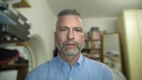 Owen Espley A close-up portrait shows Owen Espley with grey hair and beard facing the camera indoors, wearing a light blue button-up shirt. The background is softly blurred, with shelves holding books and household items on one side and what appears to be a wardrobe or mirror on the other. The lighting is even and natural, with neutral tones throughout the room, creating a calm, domestic setting.