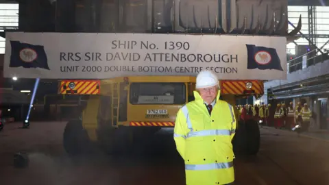 PA Media Sir David Attenborough, wearing a hi-vis jacket and white hard hat, standing in front of a yellow crawler vehicle that is carrying the bottom section of the ship that bears his name. The words: "Ship No. 1390 RRS Sir David Attenborough, unit 2000 double bottom centre section" are written on a banner draped across the section.