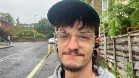 A man wearing glasses and a cap stood in the street looking into the camera. There are rain drops on his glasses.
