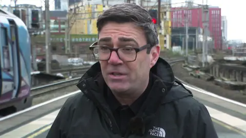 Andy Burnham trägt eine Brille und einen dunklen Mantel und steht auf einem Bahnsteig in Manchester