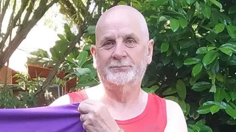 Glenn Bate Glenn Bate, a man who is standing outside in front of a green leafy bush. He is looking directly at the camera and is smiling. He is also holding up a purple T-shirt to the camera.