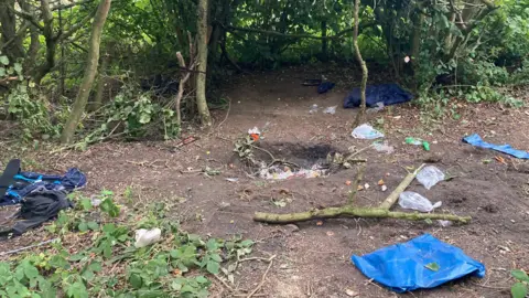Hazlerigg Parish Council An abandoned campsite where a fire had been set in a hole dug in the ground, with the surrounding area covered in strewn litter.