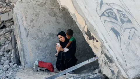 Reuters Uma mulher palestina e uma criança descansam sob os escombros de uma casa destruída em Khan Younis, no sul da Faixa de Gaza (16 de outubro de 2024)