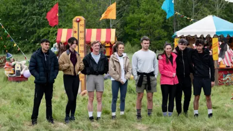 BBC/Studio Lambert/Euan Cherry/PA Wire A group of eight people who are in the Traitors standing in a made-up retro fairground, with Claudia Winkleman stood with her back to the camera and cut out of the picture.