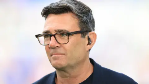 Andy Burnham, wearing an in ear monitor, glasses and a suit, looking off to the left of the picture.