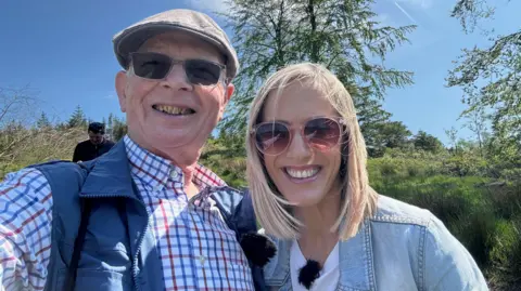 Kay Crewdson is standing next to Roger Hordley in Macclesfield Forest. They are both smiling at the camera and are wearing sunglasses. Kay is wearing a denim jacket and white T-shirt and Roger is wearing a chequered shirt and blue gilet.