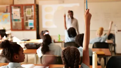 Getty Images Seven small pupils sit in a classroom with their backs to the camera. Three of them have their hands in the air. They are all wearing a light-blue polo shirt. The teacher wears a white shirt. They all sit at brown wooden desks. Their is a interactive whiteboard behind the teacher. 
