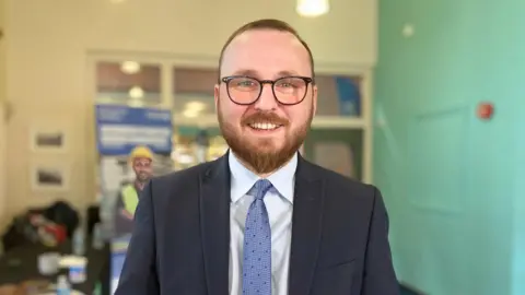 Photograph of Jack Sargeant, wearing a black suit, white shirt and blue tie. He wears black square rimmed glasses and smiles at the camera. He has a ginger-brown beard and blue eyes.