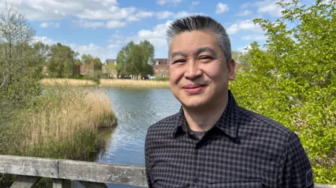 Ben Schofield/BBC Phil Yau standing outside by a lake, smiling and looking down the camera. It is a bright day and the sky is bright blue, with several puffy clouds overhead. Phil is wearing a checked shirt, worn open at the collar. He has short, neatly cut black and grey hair, which is spiked upwards. The lake behind him looks calm and flat, with trees and tall grasses along the bank. Houses and trees can be seen on the other side of the water.