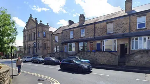 Google A street view image shows a row of houses with parked cars along the pavement. On the pavement opposite, a person is walking along a stone wall.