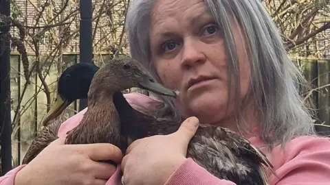 Sam poses for the camera, holding her two ducks. She has grey hair.