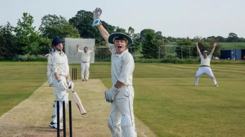 A group of teenage boys play cricket on a green. They are wearing cricket whites and holding their hands up in the air.