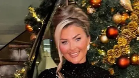 Taylor Jenkins is standing in front of a Christmas tree and smiling brightly at the camera. Her blonde hair is pulled into a bun and she is wearing a sparkly black dress and earrings.