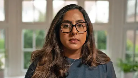 Amarjit has long brunette hair and black-rimmed glasses. She is wearing a blue jumper and a silver necklace.