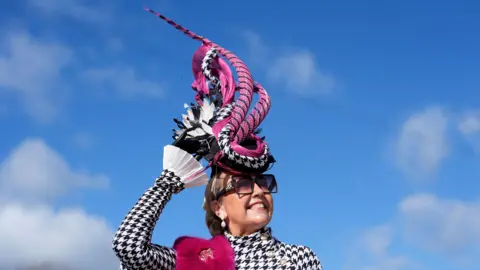 PA Media A woman with a black and white checked jacket holds onto a dramatic piece of headgear at the second day of the 2026 Cheltenham Festival. The hat is made of long, curling pieces of black and purple material
