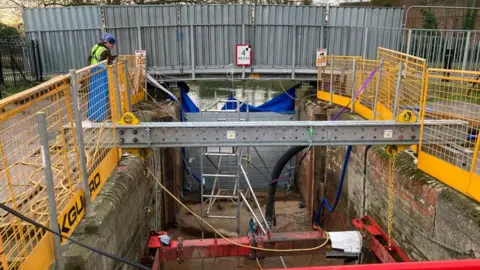 Alex Pope/BBC Work being carried out on a lock, with two men at one side, machinery bellow it, metal barriers, metal frames, a bridge and a metal shutter blocking off the other end. 