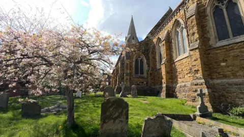 Pink blossom trees bloom within the graveyard section of the church. Gravestones and crosses sit on the green grass while the grand church stands in the background.