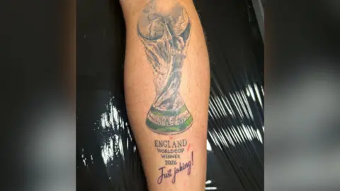 BBC Large tattoo on a man's leg. It is a trophy with a caption reading: 'England World Cup winners 2026.' with a line underneath reading: 'Just joking!'.