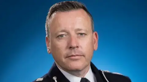 A close-up photo of the acting chief constable of West Midlands Police. He wears a black police uniform and stands in front of a blue background, looking into the camera.
