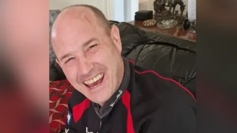 South Wales Police Martin Steel laughing at the camera in a black jersey with red detailing. He is a bald white middle aged man