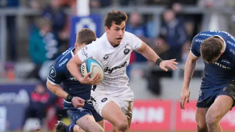 Neil Le Roux runs with the ball in his right arm as a Sale player chases behind him and another stands with his head down to his left