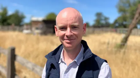 A man in a white shirt and blue gilet stands in front of a field of long grass, with miniature donkeys roaming