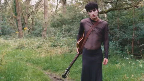 Jules Moskovtchenko A promo photo of Jacob standing in a forest with an acoustic guitar hanging from their side. They have black hair and black eye makeup. They are wearing a brown, see-through sheer top and a long black skirt.