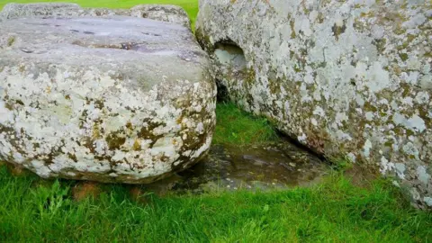 Nick Pearce Two large stones lying on grass, with one partially buried underneath.