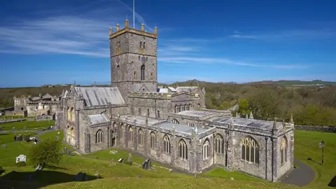 An exterior view of St Davids Cathedral. A large stone building with a square tower, it is surrounded by a grassy churchyard.