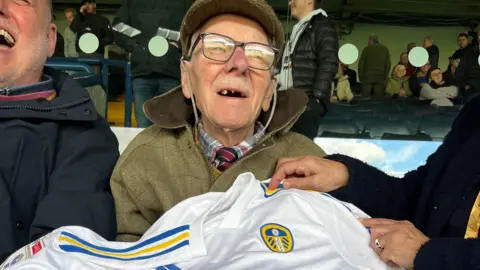 BBC/ Olivia Richwald An elderly man, wearing a tweed jacket and glasses, sits in the stands of a football ground and holds a white and blue Leeds United shirt.