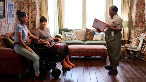 BBC / Roughcut TV / Adam Lawrence A woman in green leggings and a blue top sits on a sofa next to a woman using a wheelchair, who is wearing a shimmery top and purple skirt. On their right is a woman in green trousers and a beige shirt, holding a clip board.