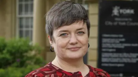 City of York Council An older woman with short grey hair and blue eyes looks at the camera. She has black hooped earrings and wears a red patterned blouse.