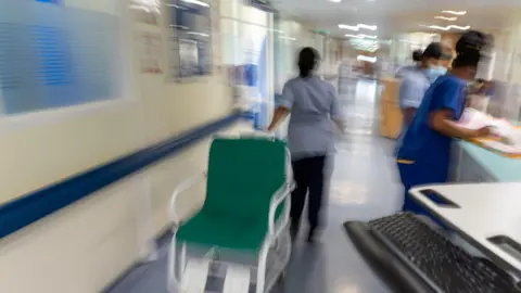 A stock image of NHS workers in a hospital corridor. They are wearing blue scrubs. One is pulling a wheelchair while two are looking at paperwork on a reception desk.