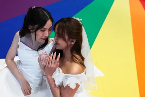 Chalinee Thirasupa/REUTERS Natnicha Somparn, 31, and Sorsomrudee Thippayatum, 32, pose for a picture as they celebrate after receiving official marriage certificates, as the country's marriage equality bill comes into effect following a royal endorsement in Bangkok, Thailand, January 23, 2025. The are wearing white dresses and are standing on a rainbow coloured painted floor.