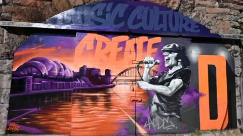 Gateshead Council A brightly coloured graffiti artwork on a railway arch door. The painting depicts AC/DC singer Brian Johnson, the Tyne bridge and has the word 'Create' written across it. Opposite Johnson is the distinctive shape of the Glasshouse centre in Gateshead.