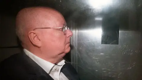 Peter Murrell, who is bald with glasses, photographed in profile in the back of a van. He is wearing a white shirt and black suit jacket, and is illuminated with a flash 
