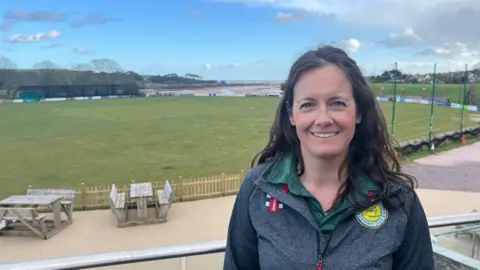 Emma Cooling in a green polo shirt and a grey zip-up top with the Budleigh Salterton Cricket Club badge in yellow and white - the lower cricket pitch is behind her with a distant view to the sea.