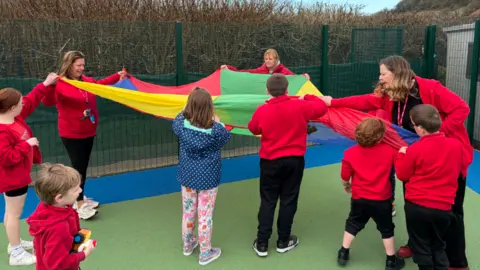 Children in red school uniform holding a multi-coloured parachute canopy with staff members