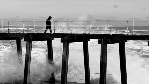 A figure walks along a pier above breaking waves