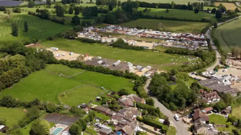 Ant Saddington/BBC Aerial view of caravans and newly developed plots at a traveller site near open countryside, with nearby homes and a larger established site in the distance.