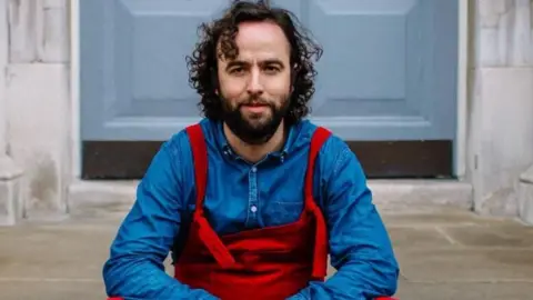 Daf James looking at the camera. He is wearing red dunagrees and a blue long sleeved top.