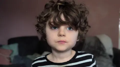 Aimee-Leigh Mahoney A six-year-old boy with curly brown hair and a black and white striped top pictured in a room