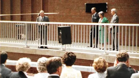 Trotman family The Duke of Edinburgh centre right stands next to a curtain covering a plaque. He has a woman and man next to him, while on the right another man is in front of a microphone on a stand. They are all on a raised walkway with white railings in front. Standing with their backs to the camera listening to the speeches are a crowd of people