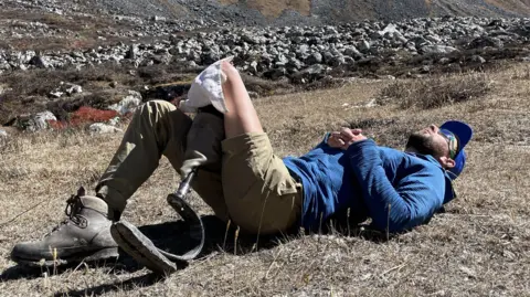 Rich Potter/Rich Davies One of the men lying on his back on grass in front of a rocky area. He is wearing a blue cap and sunglasses and has his hands clasped together on his stomach,
On his right leg is a long light-brown trouser and a black boot. The lower half of his left leg is artificial. He wears a light brown short on his left left. His bare left thigh is revealed and he has a piece of white cloth on at the point where his artificial leg is fitting to his thigh. 