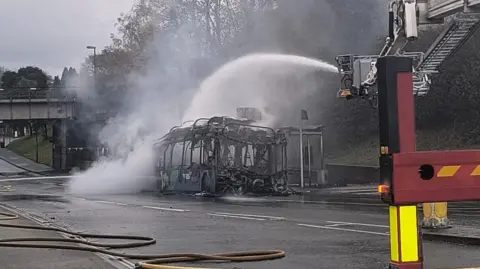 A fire tender hoses down the metal skeleton of a burnt out bus in Crawley.