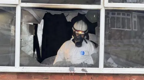 A man in an all white hazard suite and wearing a mask with a long snout over a hard hat with a lamp at the front. He is pictured behind a broken window. the room inside looks very dark