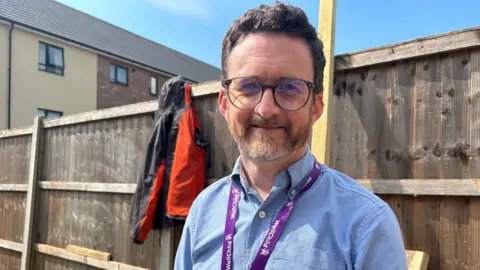 Matt smiles at the camera with a fence behind him. He wears a blue shirt and glasses and around his neck is a WellChild lanyard.
